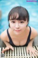 A woman in a black bathing suit leaning on a wooden bench by a pool.